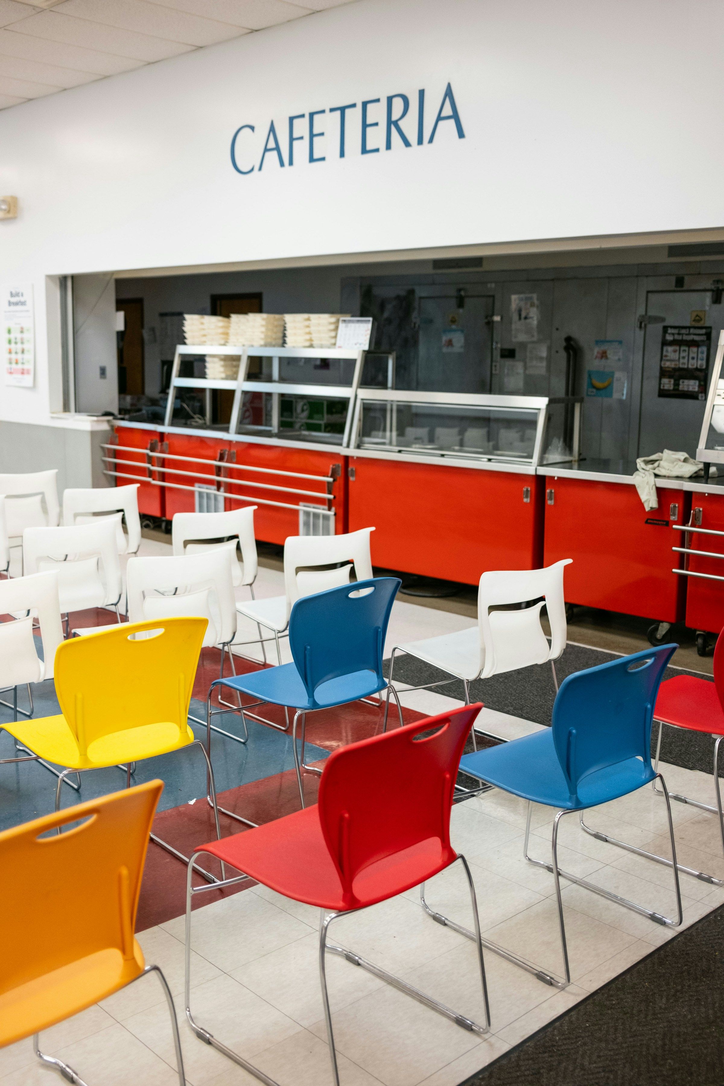 An empty school cafeteria with red serving counters and colorful stacking chairs